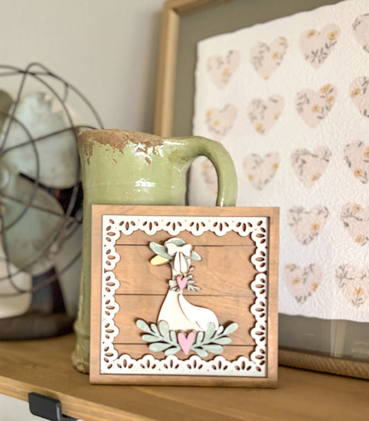 A decorative square wooden sign featuring a white goose with pastel green and pink accents, set against a rustic wood plank background with a white scalloped border. The sign is styled on a wooden shelf next to a green ceramic pitcher and a heart-patterned textile.